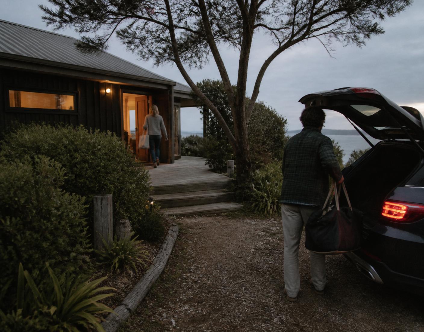 Travellers arriving at a coastal holiday house at dusk, warm light through the open door
