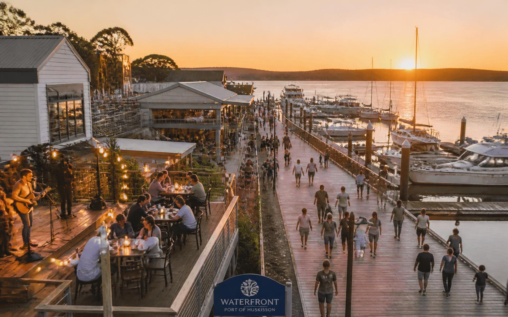 A regional waterfront precinct at golden hour with people dining outdoors, live music and families walking the boardwalk