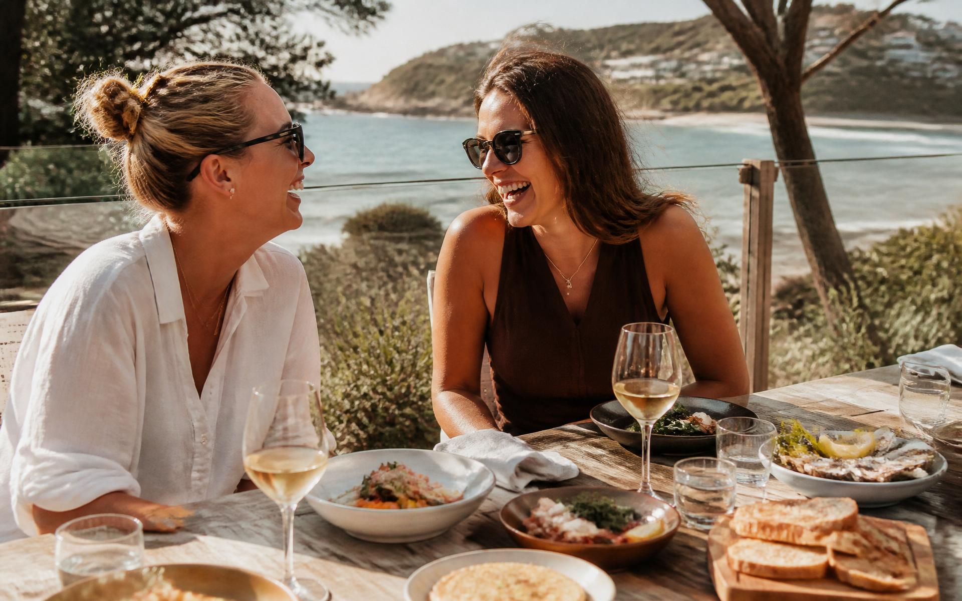 Two friends laughing over a long lunch at a coastal café