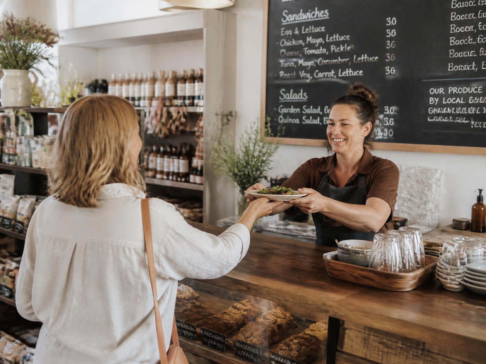 A local business owner handing a plate across the counter while visitors browse shelves of local produce