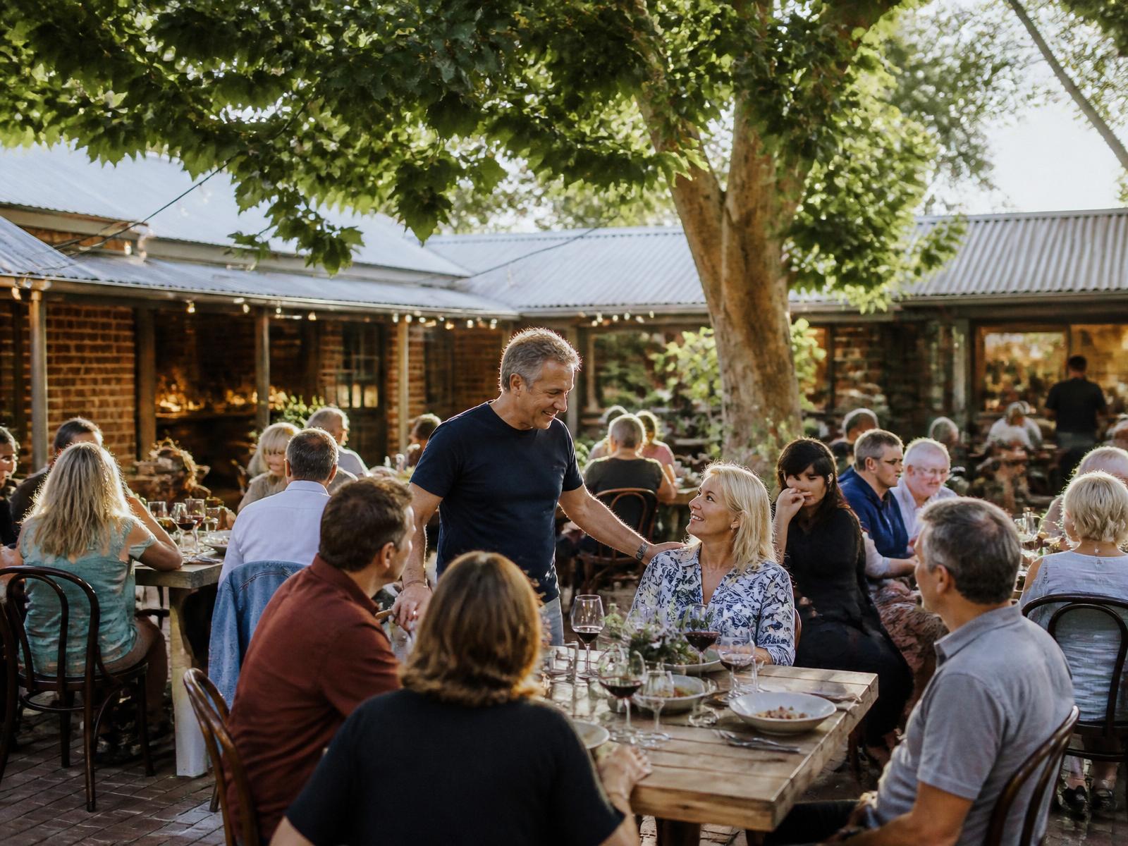A busy restaurant courtyard under string lights with full tables of guests while the owner talks with a table
